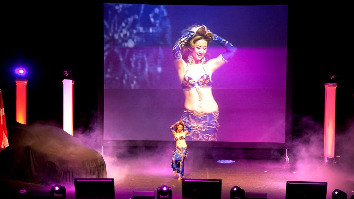 Performer on stage against a full-width projection screen, haze in the beams